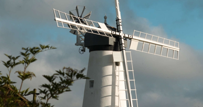 A white windmill with large sails stands against a partly cloudy sky.